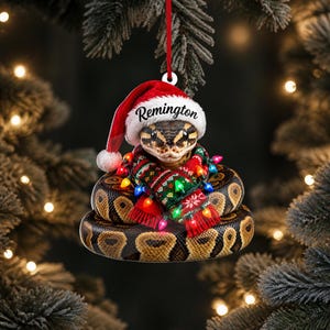 May include: A flat Christmas tree ornament featuring a coiled ball python wearing a red and white Santa hat with the name Remington printed on the brim. The snake is wrapped in a red, green, and white patterned scarf and colorful string lights.