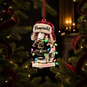 Könnte beinhalten: Ein weißes Mikroskop als Weihnachtsbaumschmuck, verziert mit einer Lichterkette aus bunten Weihnachtslichtern. Das Mikroskop trägt eine rot-schwarz karierte Trappermütze mit dem Namen Pumpernickel in schwarzer Schrift und hängt an einem roten Band.