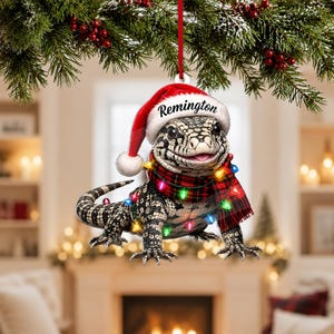May include: A flat, hanging ornament featuring a black and white lizard wearing a red Santa hat with the name Remington printed on the brim. The lizard is wrapped in a red and black plaid scarf and a string of colorful, glowing Christmas lights.