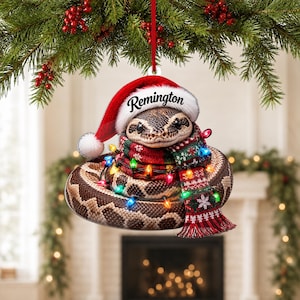 May include: A flat, circular Christmas ornament featuring a brown and tan patterned snake wearing a red and white Santa hat with the name Remington printed on the brim. The snake is wrapped in a red, green, and white patterned scarf and string lights.