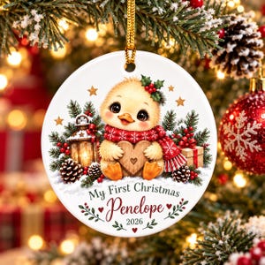 Puede incluir: Un adorno navideño redondo de cerámica blanca con un pollito amarillo que lleva una bufanda roja y sostiene un corazón de madera. El diseño incluye una linterna, ramas de pino y el texto My First Christmas Penelope 2026.