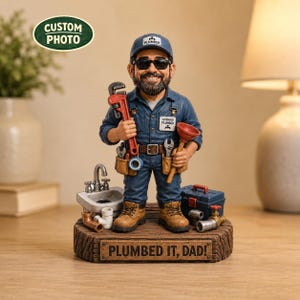 May include: A figurine of a bearded plumber wearing a blue uniform, cap, and sunglasses, holding a red pipe wrench and a plunger. The figure stands on a wooden base with the text PLUMBED IT, DAD! and includes a sink, toolbox, and pipe fittings.