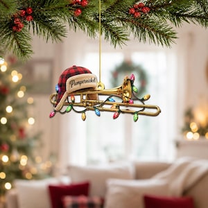 Puede incluir: Un adorno de trombón dorado con un gorro de trampero a cuadros rojos y negros con el texto Pumpernickel impreso en la parte delantera. El instrumento está envuelto en una cadena de luces navideñas de colores y cuelga de un cordón dorado.