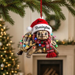 Op de afbeelding: Een hangende kerstversiering in de vorm van een schildpad met een rood-witte kerstmuts waarop de naam Remington staat gedrukt. De schildpad is omwikkeld met een snoer van gekleurde kerstverlichting en draagt een rood-groene sjaal met patronen.