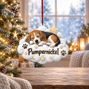 Puede incluir: Un adorno colgante en forma de nube blanca que presenta un cachorro de beagle durmiendo con pelaje marrón, negro y blanco. El adorno muestra el nombre Pumpernickel en letra marrón oscuro, acentuado con pequeñas estrellas doradas y huellas marrones.