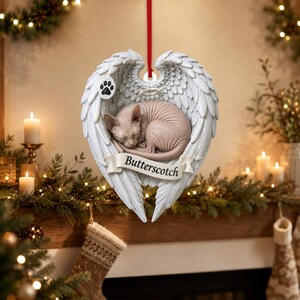 Op de afbeelding: Een hangend ornament met een lichtroze haarloze kat die opgerold in een paar witte gevederde engelvleugels ligt. Een klein zwart pootafdruk-icoon staat op de linkervleugel. Een witte banner aan de onderkant toont de naam Butterscotch.