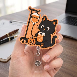 May include: A glittery badge reel featuring a black cat drinking an orange iced beverage from a cup. Next to the cat is a small jack-o'-lantern and an IV pole with an orange fluid bag labeled PUMPKIN. The badge reel has a clear plastic strap.