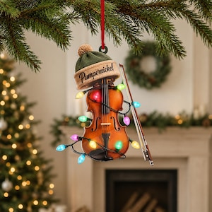 May include: A violin Christmas ornament featuring a brown wooden violin with a bow, wrapped in a string of multicoloured LED lights. The violin is topped with a green knit beanie hat that has the name Pumpernickel printed in black script.