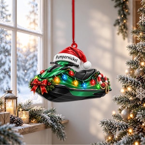 Puede incluir: Un adorno navideño de moto acuática verde colgado de una cinta roja. La moto acuática lleva un gorro de Papá Noel con el texto Pumpernickel, una cadena de luces navideñas de colores, un lazo rojo y un pequeño salvavidas en el lateral.