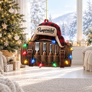 May include: A hanging ornament featuring a red and black buffalo plaid trapper hat with the word Pumpernickel printed on the front, resting atop a brown leather tool roll containing four metal chisels, wrapped in a string of colorful lights.