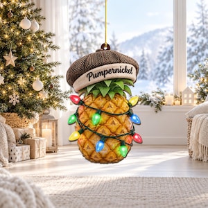 Op de afbeelding: Een hangend ornament in de vorm van een ananas met een bruine visgraat pet met de tekst Pumpernickel op de rand. De ananas is omwikkeld met een lichtsnoer met kleurrijke rode, groene, blauwe en gele kerstverlichting.