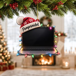 May include: A flat, hanging ornament shaped like a laptop computer. The laptop is decorated with a string of colourful Christmas lights and features a red and white knit winter hat with a pom-pom and the name Pumpernickel printed on the brim.