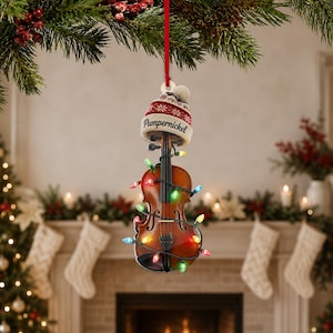 May include: A violin-shaped Christmas ornament hanging from a red ribbon. The violin is wrapped in a string of multicoloured miniature lights and topped with a cream and red knit beanie featuring the word Pumpernickel in black script lettering.