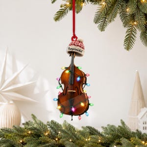May include: A brown wooden double bass Christmas ornament hanging from a red ribbon. The instrument is wrapped in a string of multicolored LED lights and wears a small red and white knit beanie cap with the word Pumpernickel printed on it.