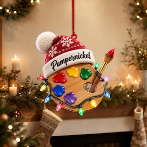 May include: A wooden artist palette ornament featuring a red knit hat with a white pom-pom and the text Pumpernickel. The palette has five colourful paint blobs, a paintbrush, and is wrapped in a string of multicoloured Christmas lights.