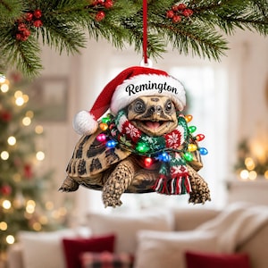 Op de afbeelding: Een hangende kerstversiering met een schildpad die een rood-witte kerstmuts draagt met de naam Remington op de rand gedrukt. De schildpad is omwikkeld met een groen, rood en wit patroonsjaaltje en een snoer met kleurrijke kerstverlichting.