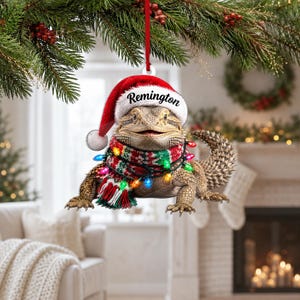 Puede incluir: Un adorno colgante plano que muestra un dragón barbudo con un gorro de Papá Noel rojo y blanco con el nombre Remington impreso en el borde. El lagarto lleva una bufanda de patrones rojos, verdes y blancos adornada con luces de colores.