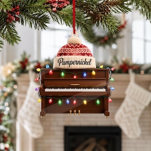 May include: A brown upright piano ornament featuring a red and white knit beanie on top with the text Pumpernickel printed on the brim. The piano is decorated with a string of colorful multi-colored Christmas lights and hangs from a red ribbon.