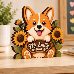 May include: A layered wooden cutout of a Corgi dog with orange and cream fur, holding a sign that reads Ms. Emily with a bone and paw print icon. The dog is flanked by two large yellow sunflowers with green leaves on both sides of the figure.