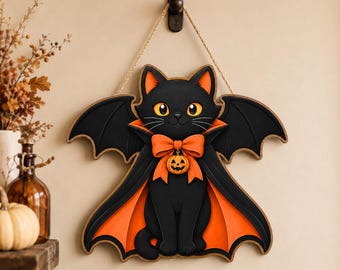 May include: A decorative wall hanging featuring a black cat with large yellow eyes, wearing a black and orange cape with bat wings. The cat has an orange bow tie with a small jack-o'-lantern charm attached, suspended by a tan rope from a hook.