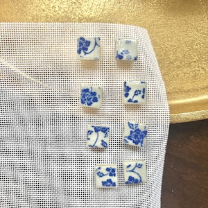 May include: Six small, square ceramic tiles with a white base and blue floral patterns are displayed. The tiles are arranged on a white mesh surface, highlighting the detailed designs and colour contrast. These tiles are likely for a craft project.