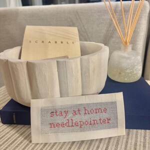 May include: A rectangular needlepoint canvas featuring the red text "stay at home needlepointer" on a white grid background. The canvas is positioned in front of a light-colored scalloped bowl, a Scrabble game box, a reed diffuser, and a dark blue book.