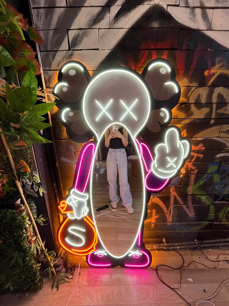 Op de afbeelding: Een neonlichtsculptuur in de vorm van een stripfiguur met een wit gezicht, zwarte oren en doorgestreepte ogen. De figuur houdt een geldbuidel met een dollarteken en een hand die de middelvinger opsteekt. De omtrek is roze.