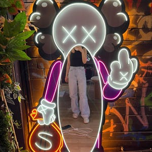 Op de afbeelding: Neon spiegel in de vorm van een stripfiguur met een wit gezicht, zwarte oren en gekruiste ogen. Het personage houdt een geldbuidel vast en steekt de middelvinger op. De spiegel heeft een paarse en witte omtrek.