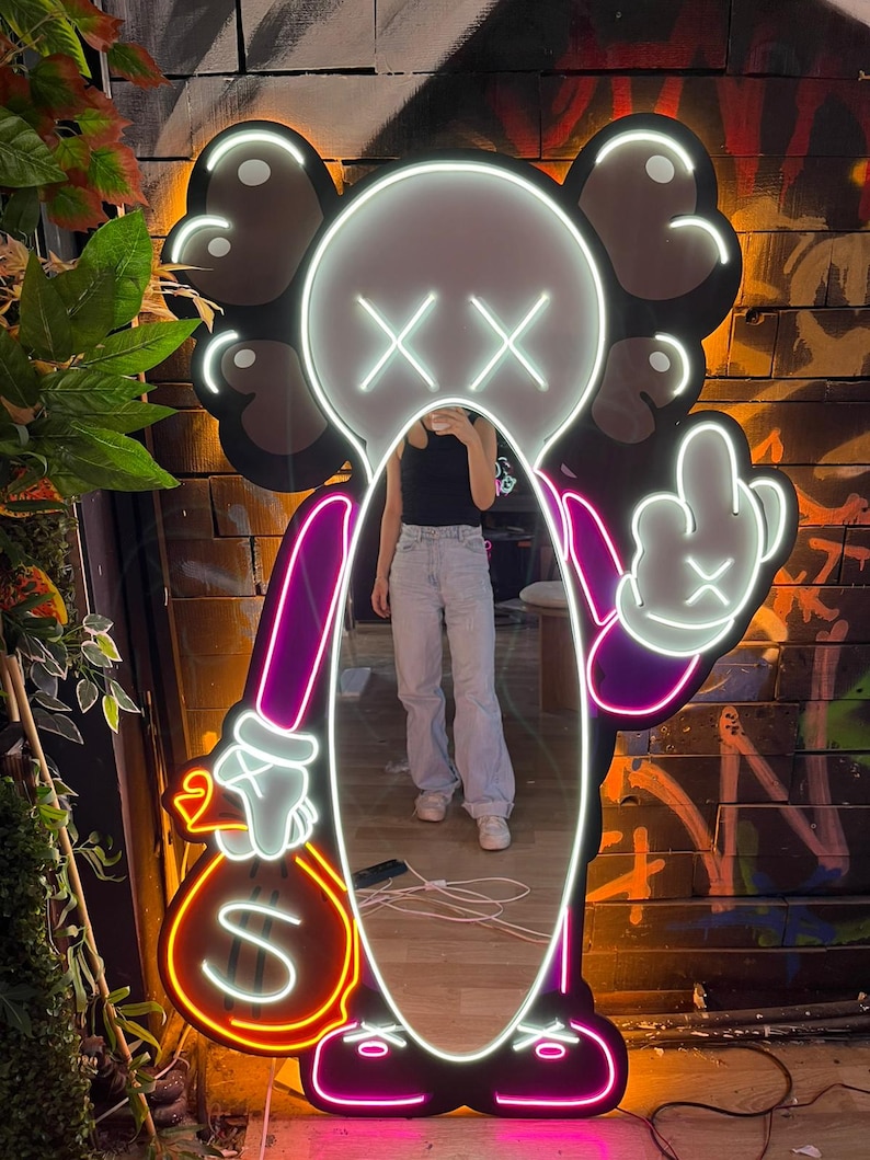 Op de afbeelding: Neon spiegel in de vorm van een stripfiguur met een wit gezicht, zwarte oren en gekruiste ogen. Het personage houdt een geldbuidel vast en steekt de middelvinger op. De spiegel heeft een paarse en witte omtrek.
