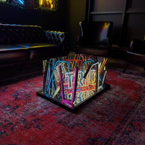Op de afbeelding: Een moderne salontafel met neonlichten die het woord "Retro" spellen in verschillende kleuren. De tafel staat op een zwarte basis, die de neonlichten weerspiegelt. De tafel staat op een rood patroon tapijt.