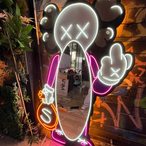 Op de afbeelding: Neonlichtsculptuur in de vorm van een stripfiguur met een spiegel als lichaam. De figuur is omlijnd in wit en roze neon, met een geldbuidel en een middelvingergebaar. De achtergrond is een bakstenen muur met graffiti.