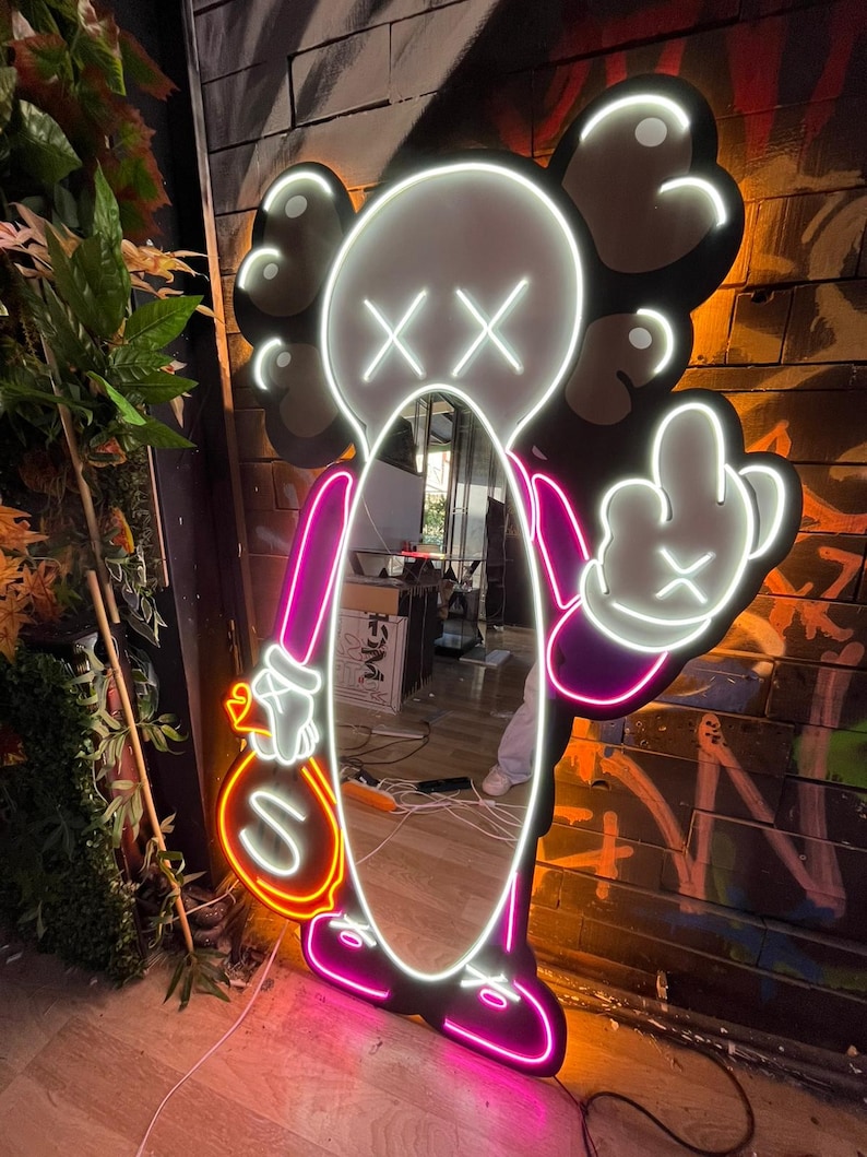 Op de afbeelding: Neonlichtsculptuur in de vorm van een stripfiguur met een spiegel als lichaam. De figuur is omlijnd in wit en roze neon, met een geldbuidel en een middelvingergebaar. De achtergrond is een bakstenen muur met graffiti.