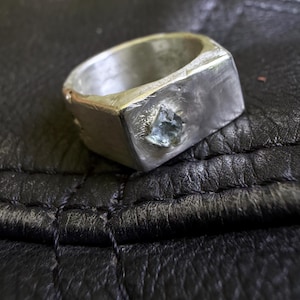 May include: A silver ring with a rectangular face and a raw, uncut blue gemstone. The ring has a textured, handcrafted appearance, with visible tool marks. The band is thick and solid, giving the ring a bold, modern look.
