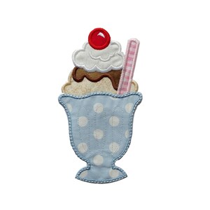 May include: A fabric applique of an ice cream sundae. The sundae features a light blue polka-dotted cup, topped with vanilla and chocolate ice cream, whipped cream, and a red cherry. A pink and white checkered straw is inserted into the sundae.