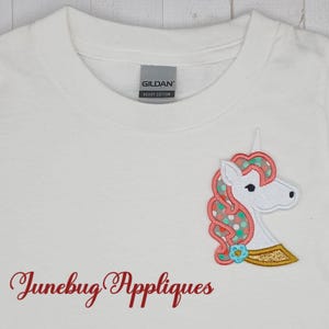 May include: White t-shirt with a pink and green unicorn applique. The applique has a gold trim and a small blue flower. The text "Junebug Appliques" is printed on the shirt.