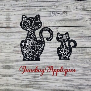 May include: Two black cat-shaped fabric appliques featuring a white spiderweb pattern throughout their bodies. The larger cat is on the left and the smaller cat is on the right. The red text Junebug Appliques is positioned below the two cat figures.