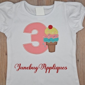 May include: White short-sleeved t-shirt with a pink number 3 and an ice cream cone applique. The ice cream cone has layers of yellow, pink, and blue ice cream with sprinkles and a cherry on top. The text "Junebug Appliques" is written in red.