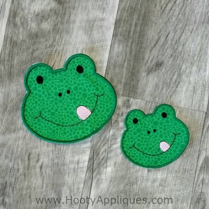 May include: Two green frog-shaped fabric appliques with a dotted texture, black embroidered eyes, a curved smile, and a small pink tongue. The larger frog is on the left and the smaller one is on the right. The text HootyAppliques.com is at the bottom.