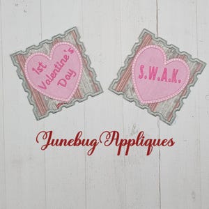 May include: Two pink felt heart-shaped appliques with embroidered text. The first applique says "1st Valentine's Day" and the second applique says "S.W.A.K.". The appliques are on a white wood background with the text "Junebug Appliques" below.