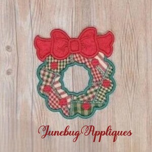 May include: A fabric applique featuring a circular Christmas wreath design with a red bow on top. The wreath is composed of patchwork squares in red, green, and white plaid patterns with small red circular accents. The text Junebug Appliques is below.