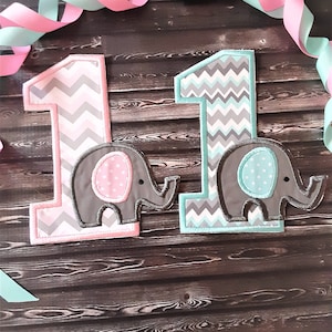 May include: Two fabric number one appliques featuring a gray and white chevron pattern. The left number is outlined in pink with a gray elephant applique featuring a pink polka dot ear. The right number is outlined in teal with a gray elephant applique featuring a teal polka dot ear.