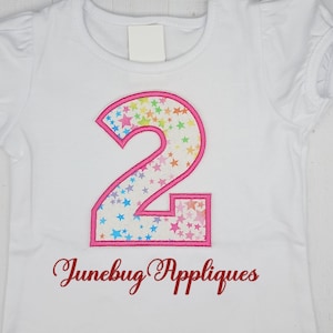 May include: White t-shirt with a pink number two applique. The number two is filled with a rainbow star pattern. The text "Junebug Appliques" is embroidered in red below the number.