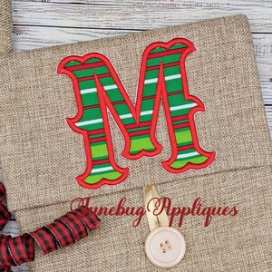 May include: A tan burlap Christmas stocking featuring a large embroidered letter M with horizontal red, green, and white stripes. The stocking includes a beige button closure and the red embroidered text Junebug Appliques.