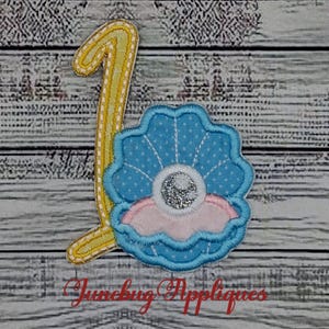 May include: A fabric applique featuring a yellow number one with white stitching next to a blue polka-dot seashell containing a pink base and a silver glittery pearl center. The text Junebug Appliques is printed in red at the bottom.