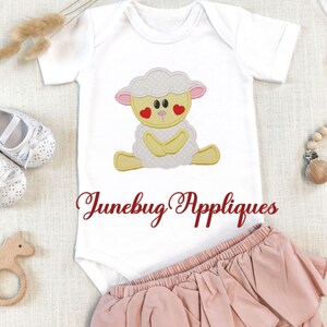 May include: White baby onesie with a yellow and white embroidered lamb with red hearts for eyes. The onesie has the text "Junebug Appliques" in red embroidery on the bottom.