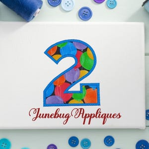 May include: A white fabric with a blue outlined number two. The number two is filled with a pattern of colorful balloons. The text "Junebug Appliques" is written in red below the number.