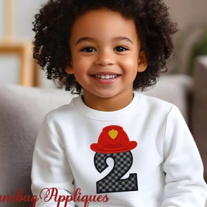 May include: A young child wearing a white sweatshirt with a black and red firefighter applique. The applique features a number "2" with a red firefighter hat on top.