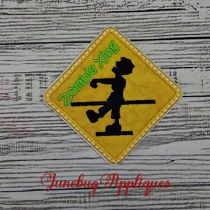 May include: A diamond-shaped yellow fabric patch featuring a black silhouette of a walking figure and the green embroidered text "Zombie Xing". The patch has a white stitched border and is designed as a novelty road sign warning graphic.