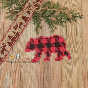 May include: A red and black buffalo plaid bear applique with a white outline. The bear is on a wooden surface with a decorative ribbon featuring trees and moose. Greenery and a bow are also present.