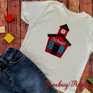 May include: White t-shirt with a red schoolhouse applique featuring blue windows and the word "School". A pair of blue denim jeans and colorful wooden blocks are also in the image.
